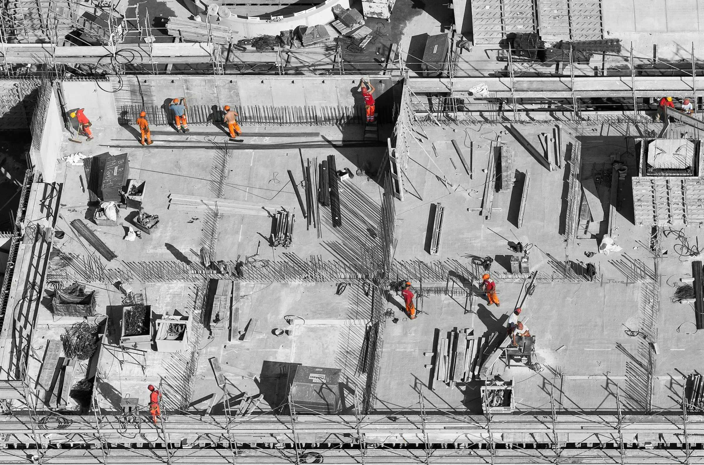 Aerial view of a concrete building site where more than a dozen workers in orange high-visibility clothing work across the deck.