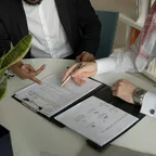 Two men at a table pointing at printed documents with a pen, one in a white thobe and red-checked shemagh