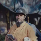 A bearded worker in a hard hat and work shirt holding a tablet on site