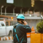 A worker in a blue hard hat and harness talking on a mobile phone at a roadside work site
