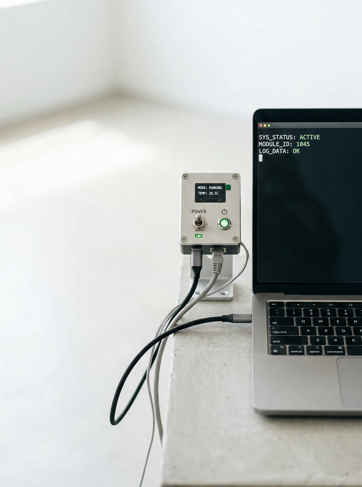 Small electronic device with screen showing 'MODE: RUNNING TEMP: 28.5C' connected by cables to a laptop displaying system status text.