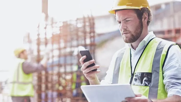 Construction worker in a yellow hard hat and safety vest using a smartphone and holding a clipboard at a building site.