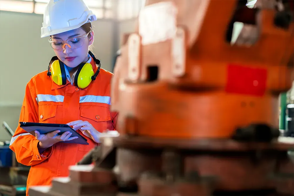 Technician in orange coveralls, safety glasses and ear defenders checking a tablet next to industrial machinery