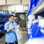 A storeroom worker in a blue uniform checking a paper list against stock on shelves of blue parts bins