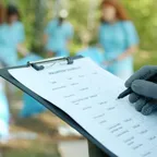 A gloved hand filling in a paper checklist on a clipboard outdoors