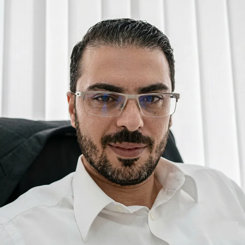 Man with dark hair, beard, and glasses wearing a white shirt sitting in front of white curtains.